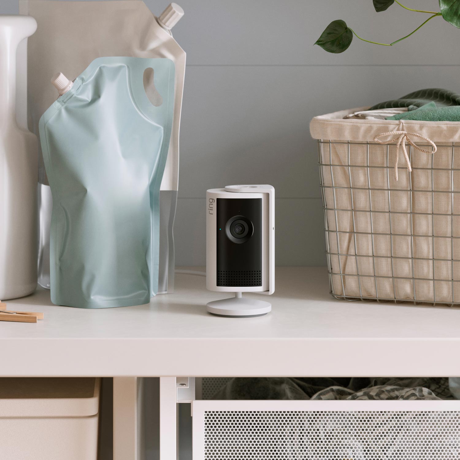 Indoor Cam Plus (Featuring Retinal 2K) - A white Indoor Cam Plus mounted on a wall next to a wooden shelf. Below the shelf is a small plant in a blue-grey pot.