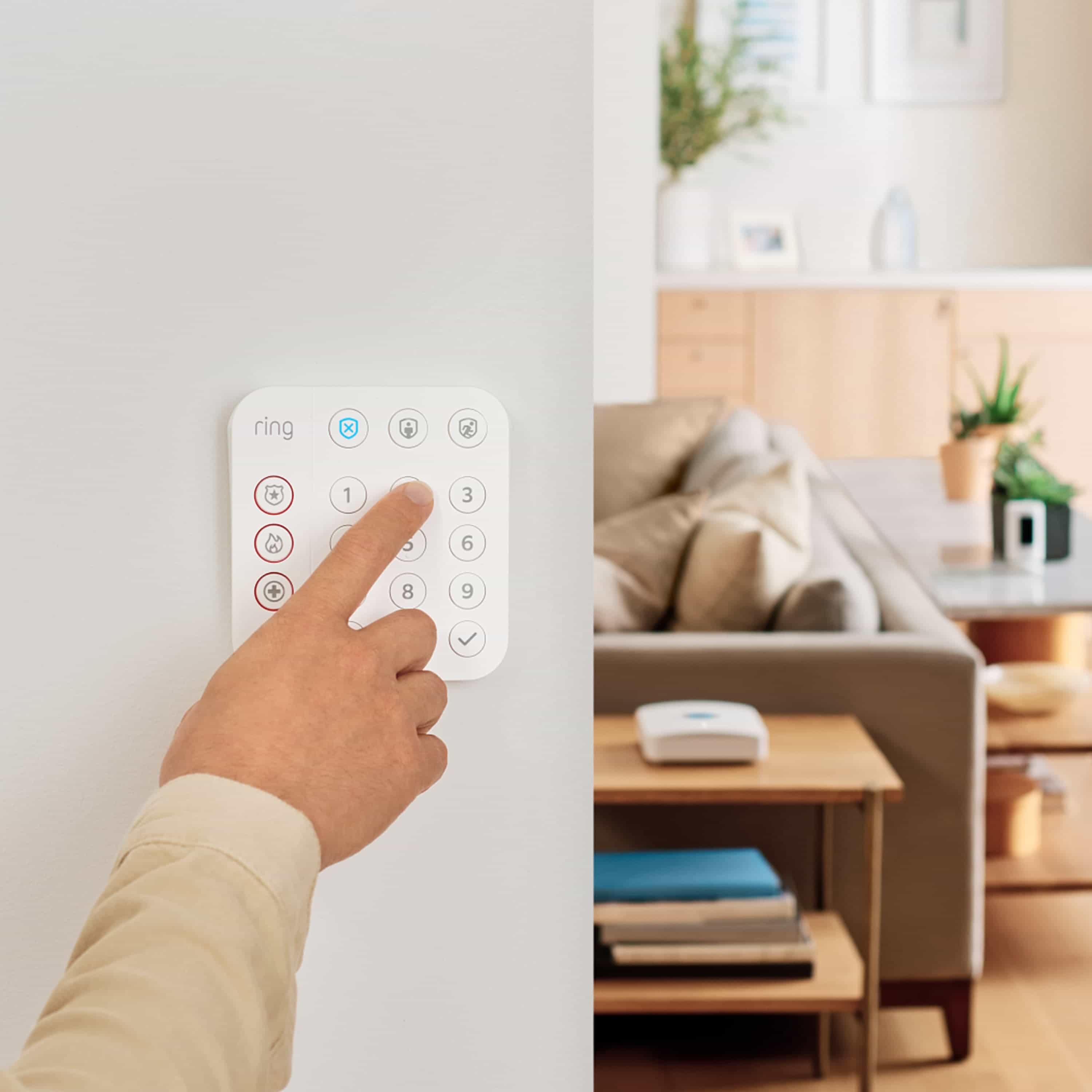 Alarm Security Kit, 10-Piece (for 2nd Generation) - Person pressing a button on a white Ring Alarm Keypad mounted on a wall, with a living room and Ring Base Station visible in the background.