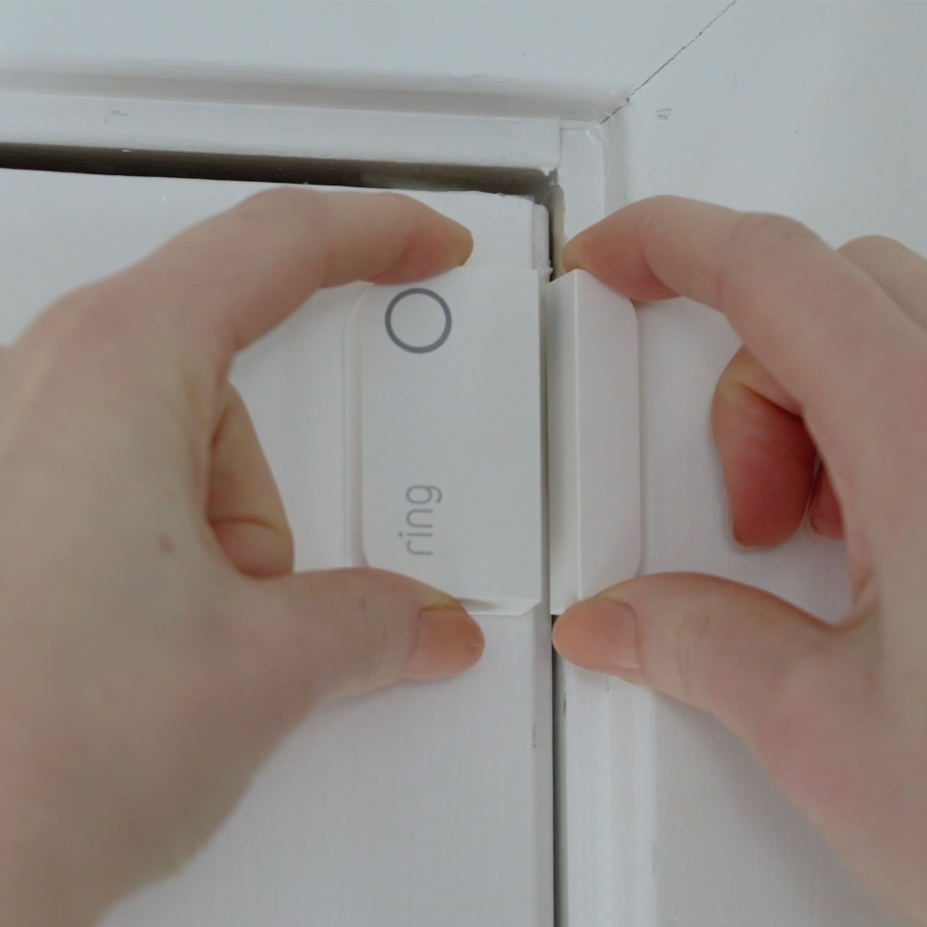 Alarm Window and Door Contact Sensor - Hands installing a white Ring Alarm Window and Door Contact Sensor on a white door frame.