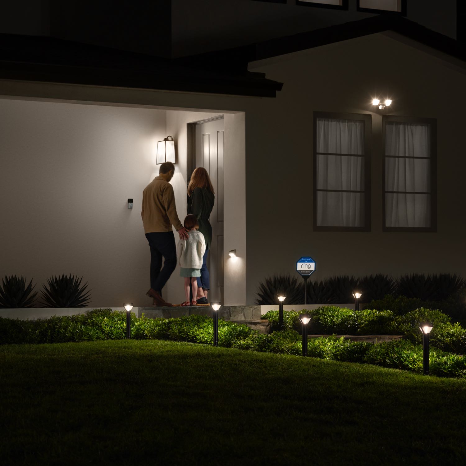 Smart Lighting Pathlight Battery - Family entering a well-lit home at night with Ring Smart Lighting Pathlights illuminating the walkway and yard.