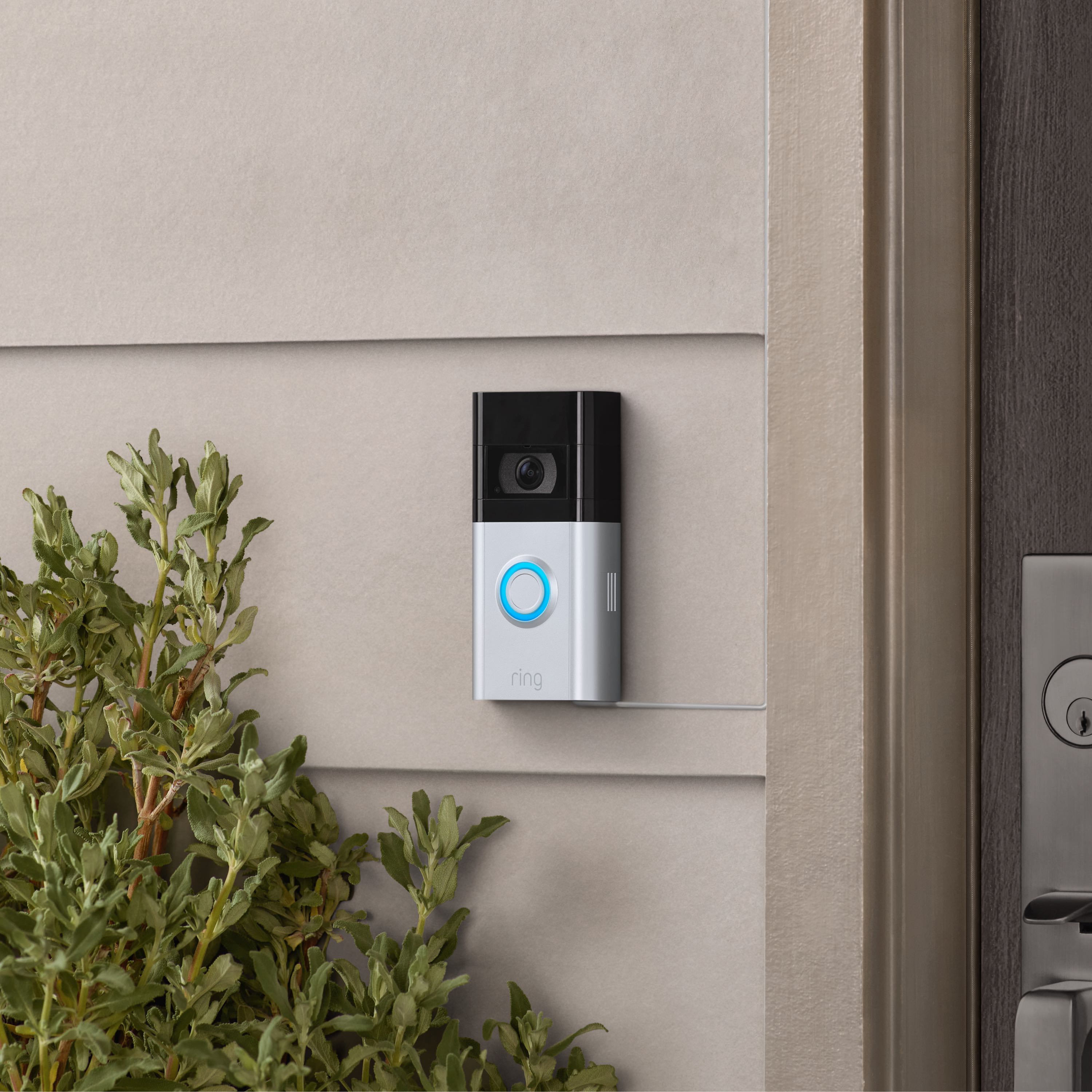 Charging Station + 2 Quick Release Battery Packs - Ring Video Doorbell with blue-lit button mounted near a door, shown next to plants.
