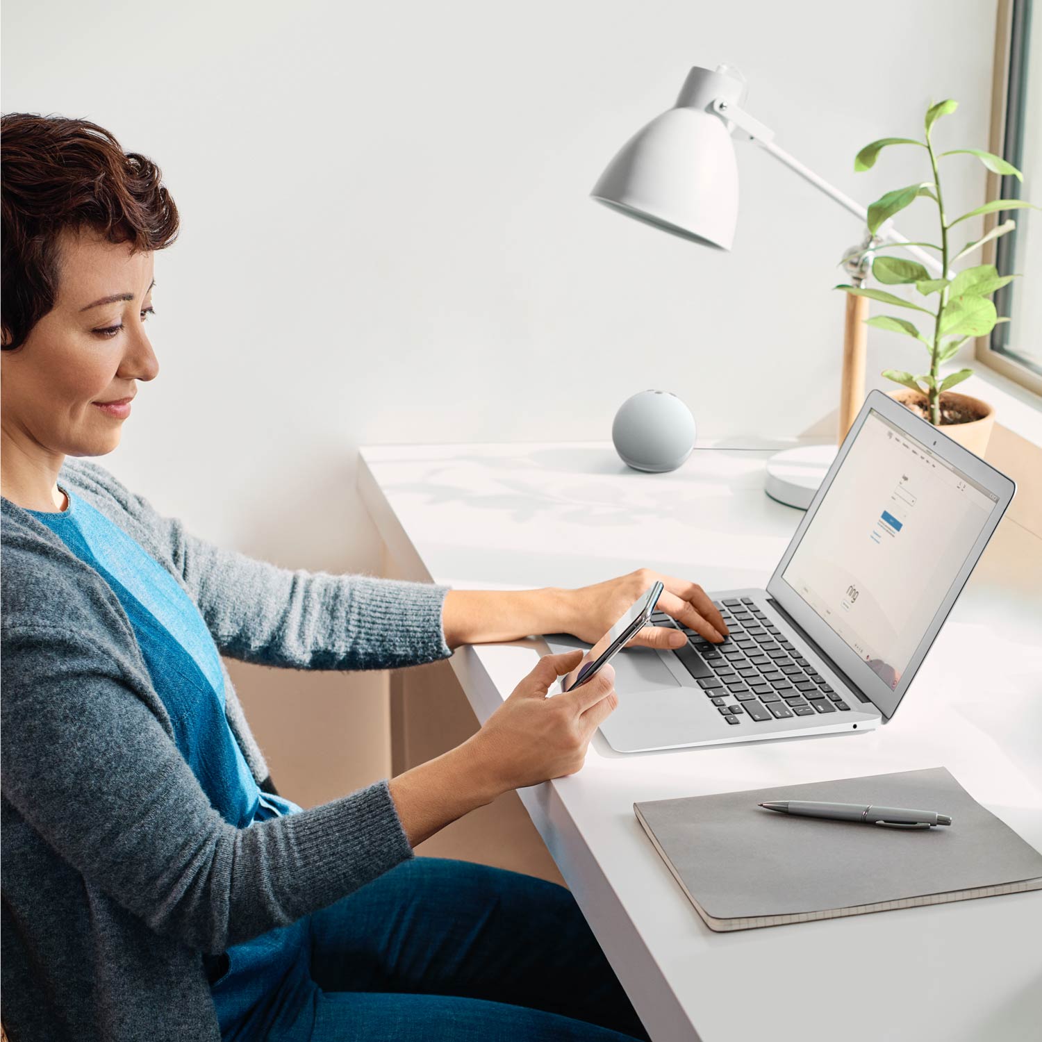 Alarm Pro Base Station (for with built-in eero Wi-Fi 6 router) - Woman sitting at white desk using a smartphone and laptop, with potted plant, desk lamp, and smart speaker nearby.