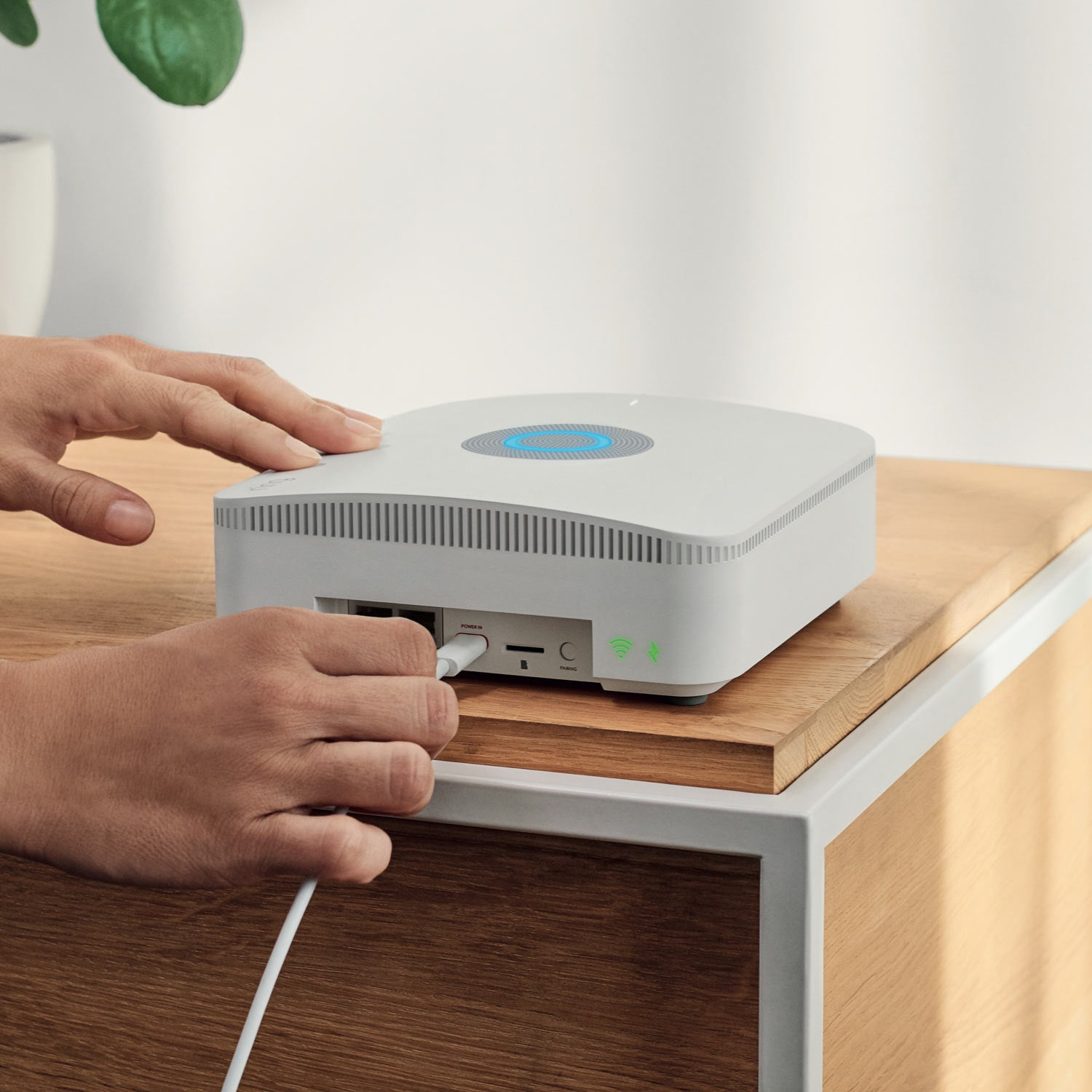 Alarm Pro Base Station (for with built-in eero Wi-Fi 6 router) - Person plugging cable into back of white Ring Alarm Pro Base Station on table, with blue light ring glowing on top.
