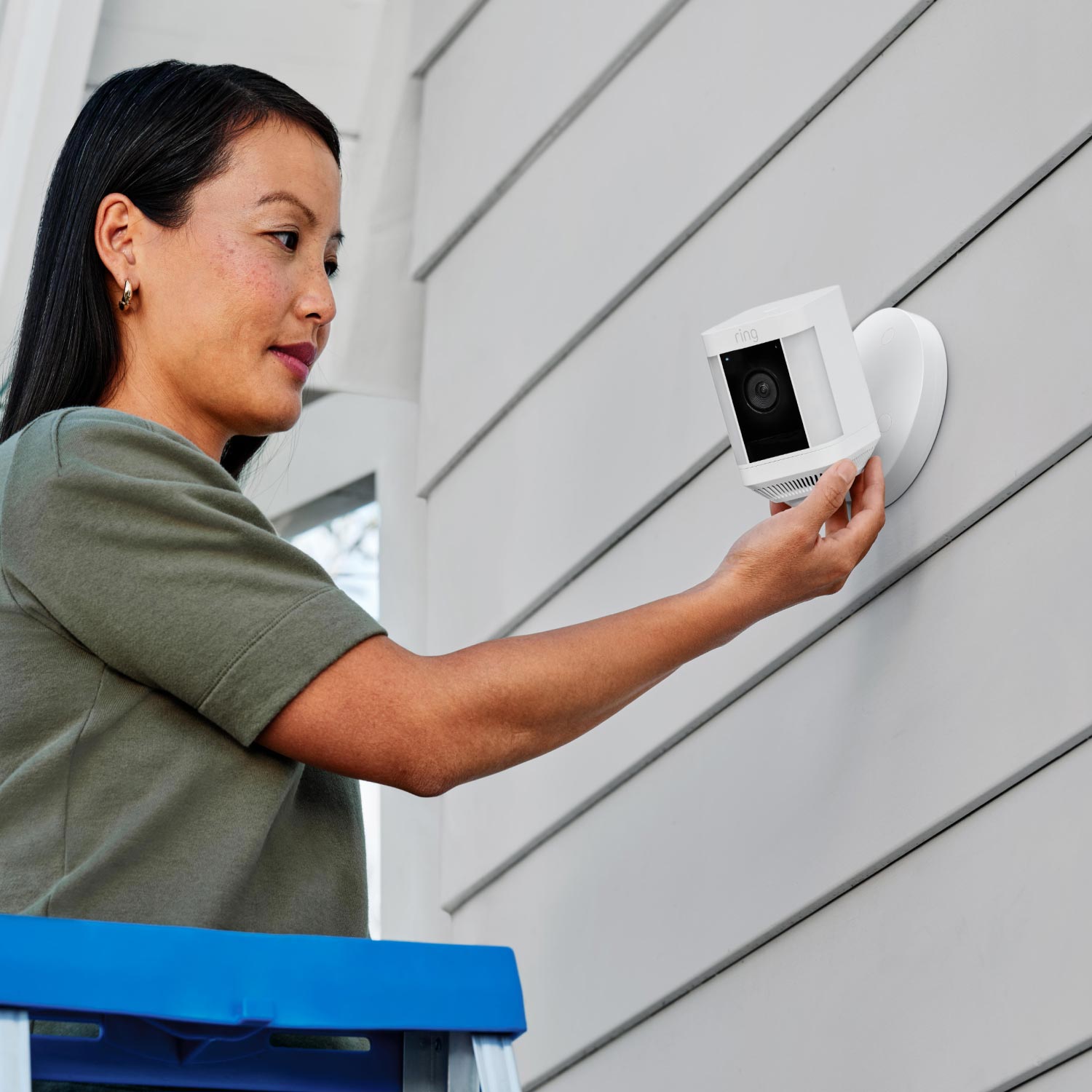 Hardwired Kit for Cameras (Outdoor Cam Plus, Spotlight Cam Plus, Spotlight Cam Pro) - Woman on a ladder installs a white Ring Spotlight Cam on a wall using the Hardwired Kit's round mounting plate.