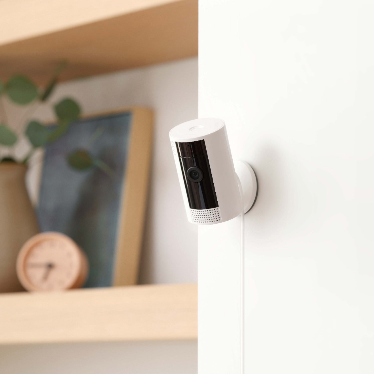 8-Piece Alarm Security Kit + Indoor Cam (2nd Gen) - White Ring Indoor Cam 2nd Gen on wall with cable visible, next to a shelf with a clock, a vase, and framed art.