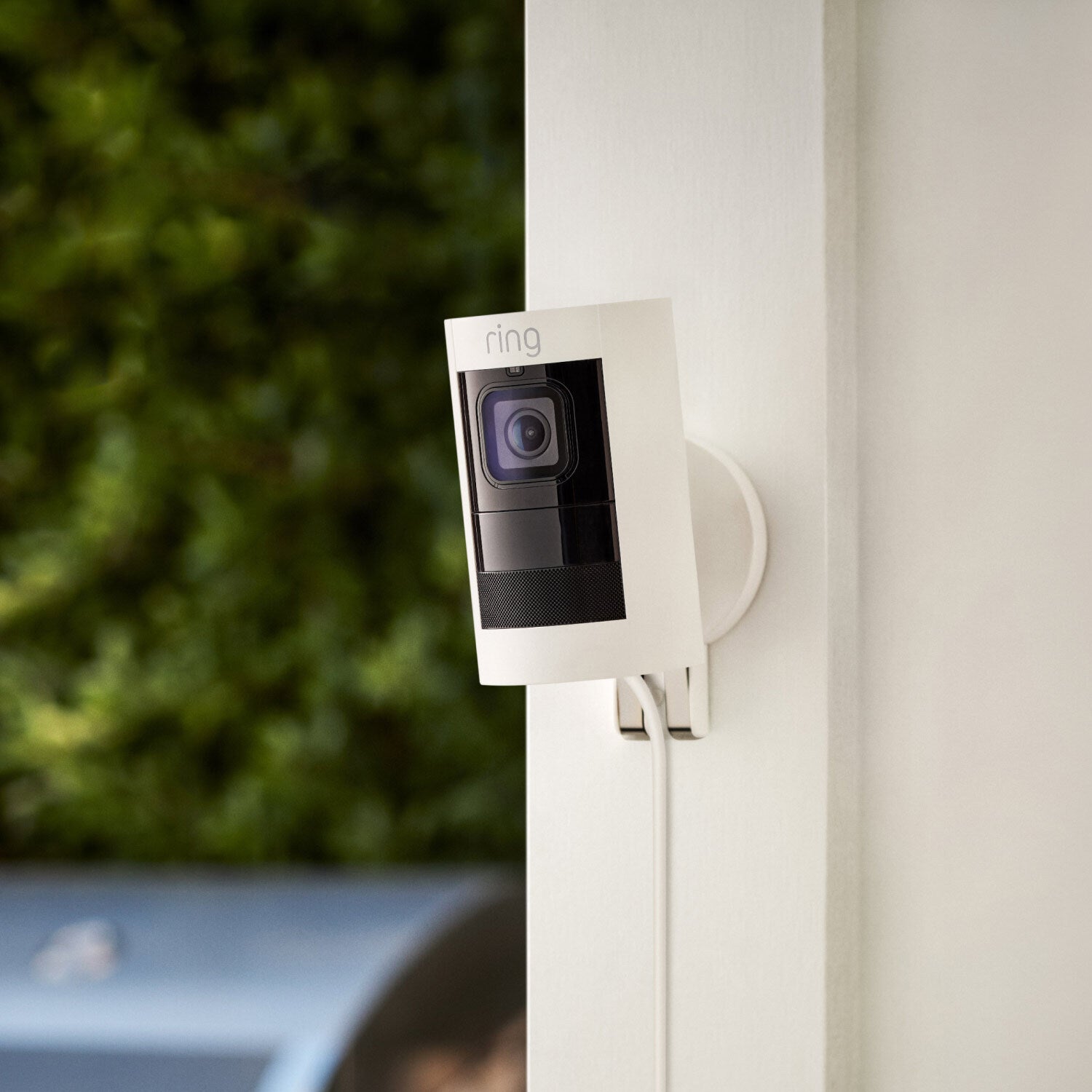 Stick Up Cam Elite with PoE Adapter - White Ring Stick Up Cam Elite with PoE Adapter mounted outdoors on a white wall, connected by PoE cable, with greenery blurred in the background. 