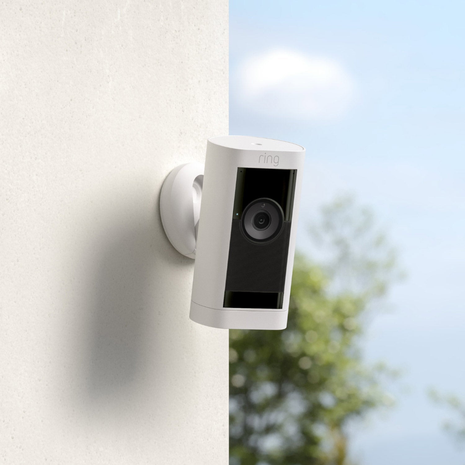 Stick Up Cam Pro (Certified Refurbished) - Battery:White Ring Stick Up Cam Pro mounted on outdoor wall, angled downward, with blue sky and tress in background.