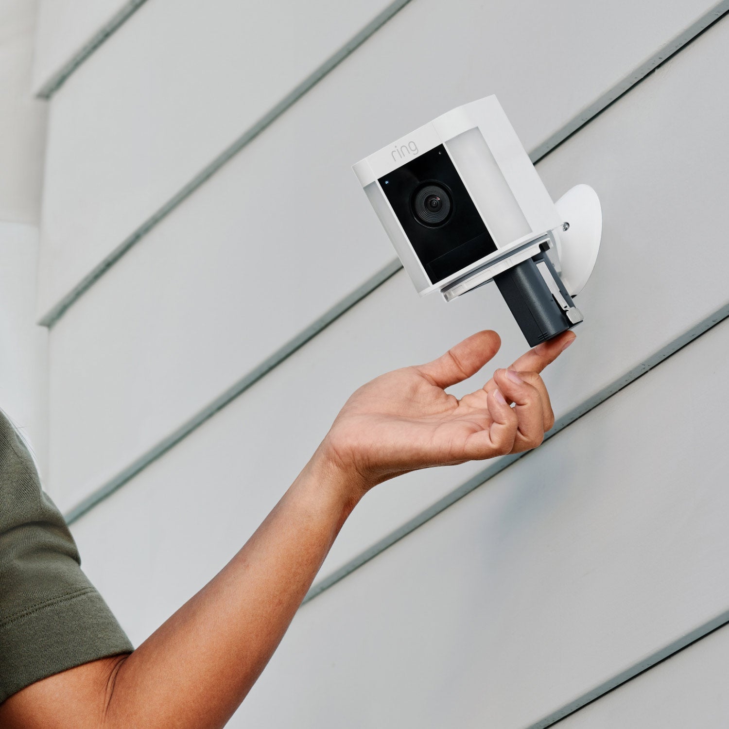Spotlight Cam Plus - Battery:Person inserting a battery into a white Spotlight Cam Plus mounted on an exterior wall with horizontal siding.