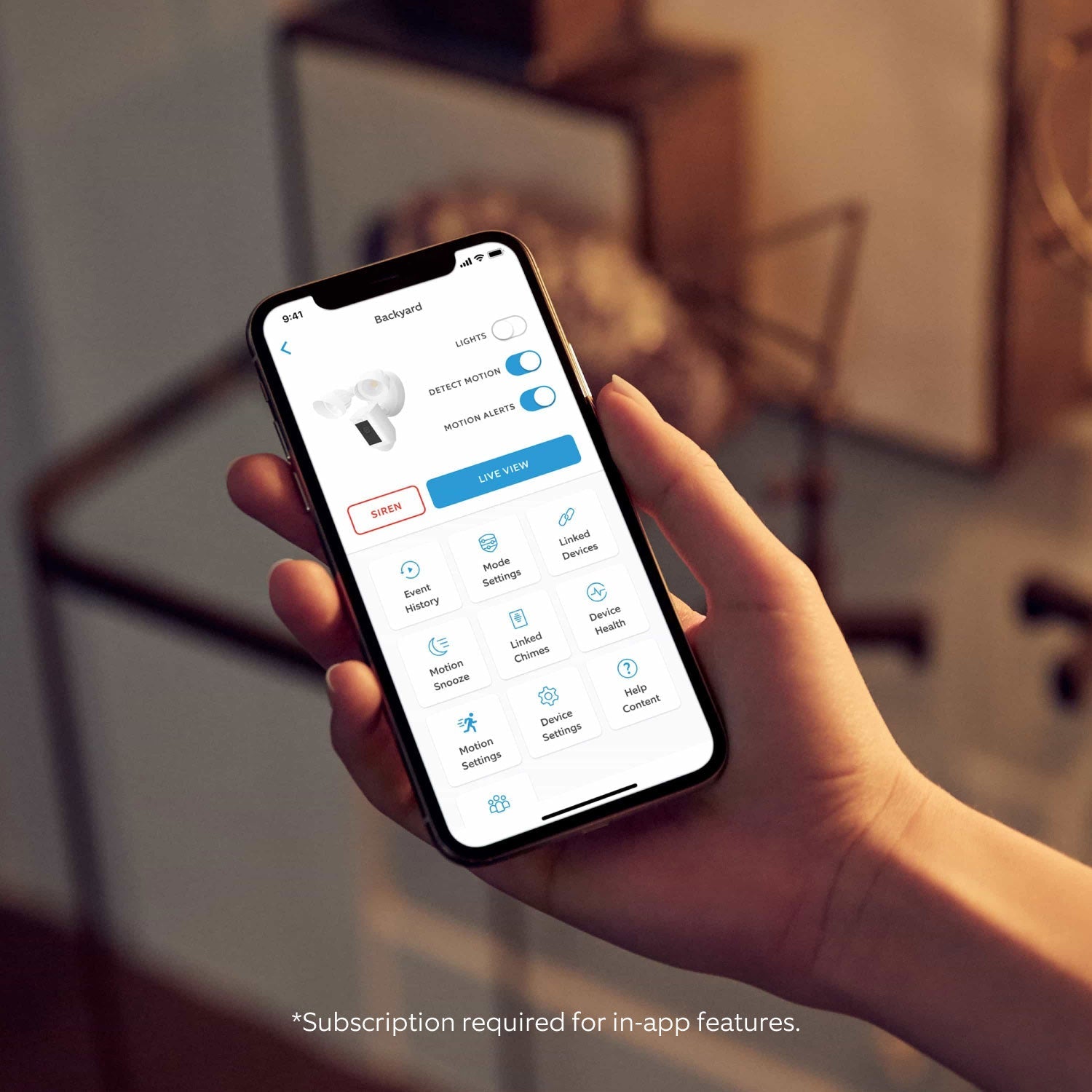 Floodlight Cam Pro (Featuring Retinal 2K) - Close-up of hand holding smartphone. Screen shows Floodlight Cam settings in Ring app. Text reads, 'Subscription required for in-app features.'