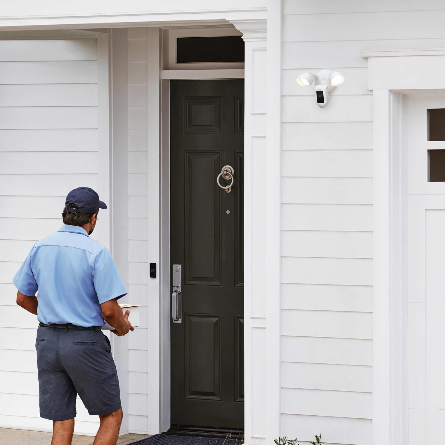 Floodlight Cam Plus - Delivery person approaches front door with package while Ring Floodlight Cam Wired Plus monitors from above.