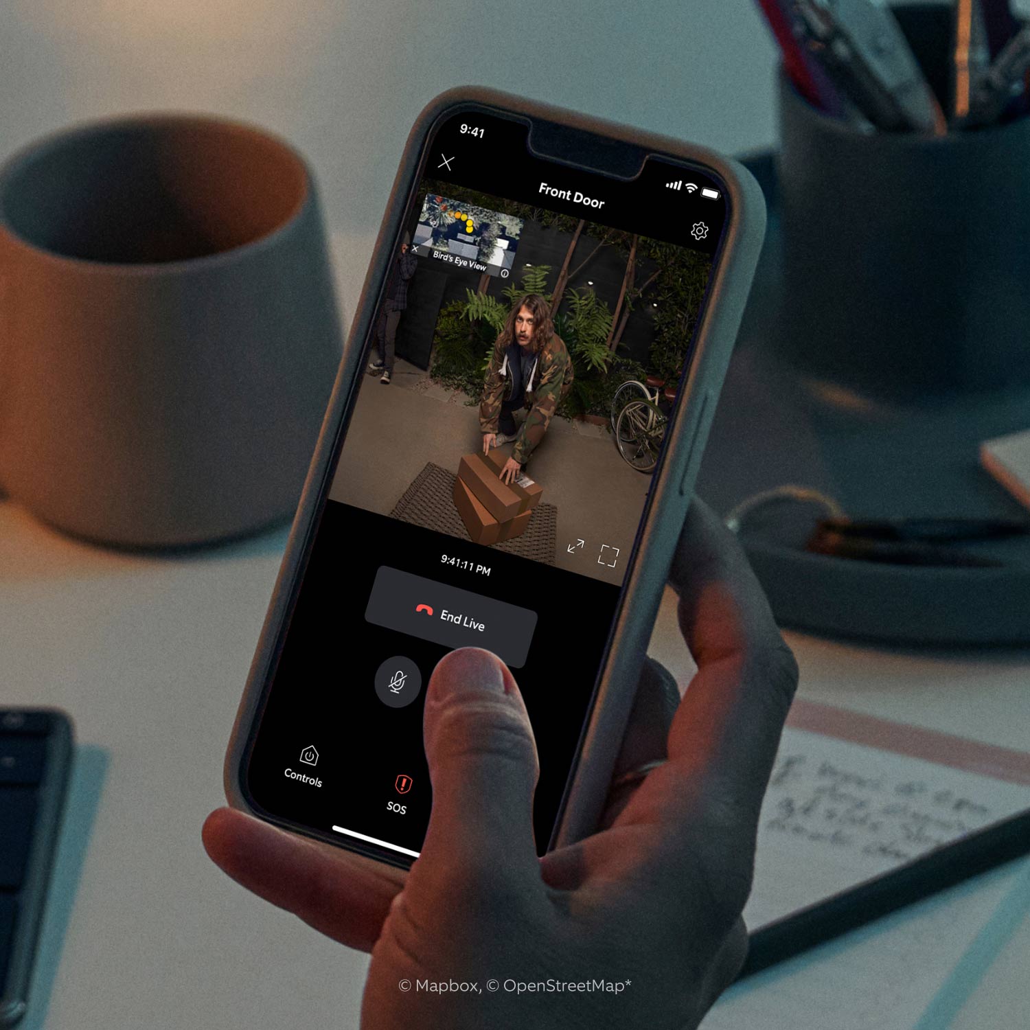 Battery Doorbell Pro - Hand holds smartphone displaying live Ring App doorbell feed of a man in hoodie picking up packages at front door.