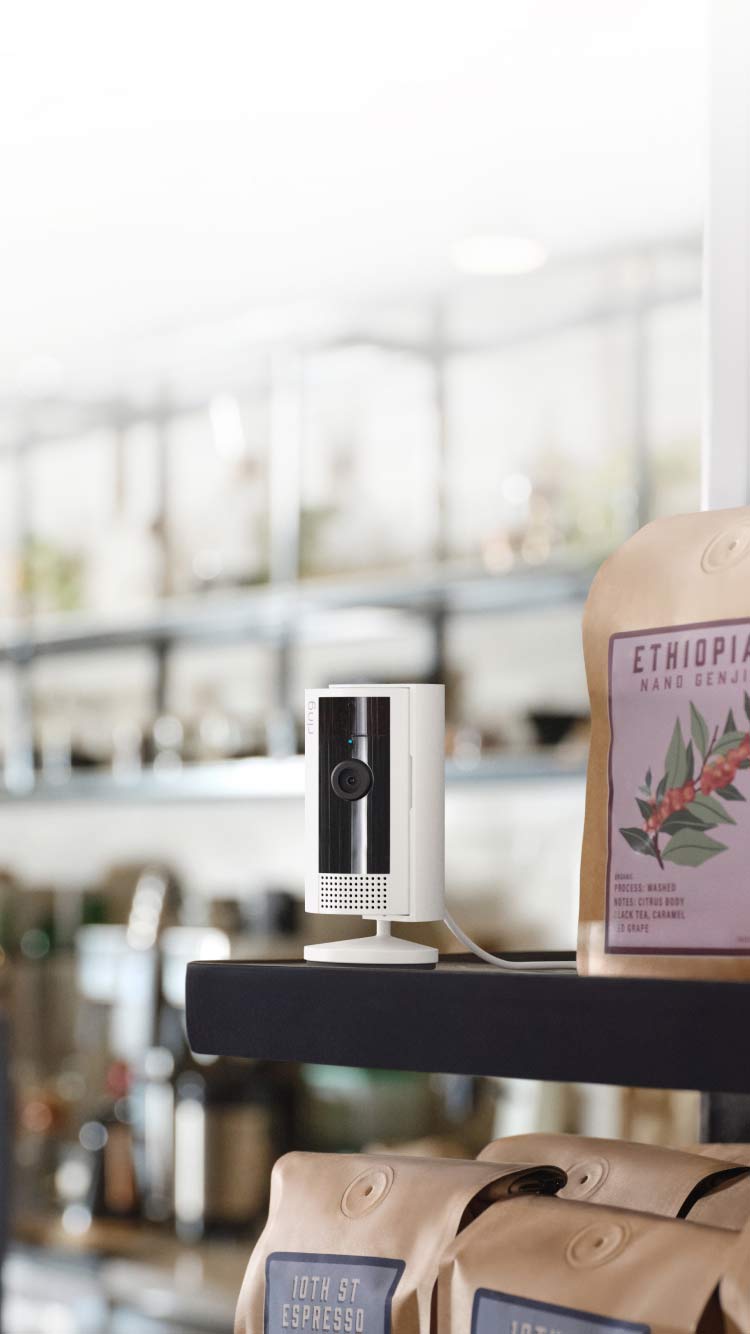 A white Ring Spotlight Cam Plus mounted and illuminated on a building next to a shop window.