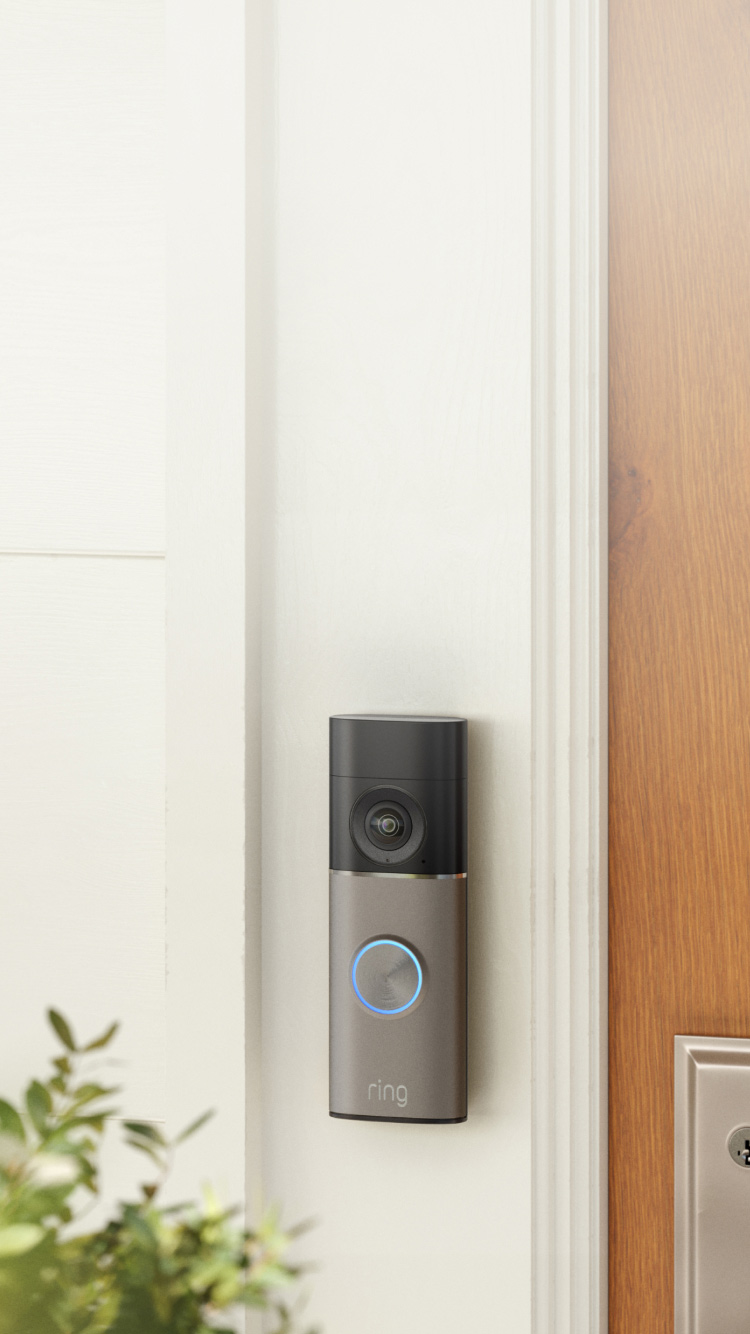 Ring video doorbell mounted on white doorframe next to wooden door, with green plants visible in foreground.