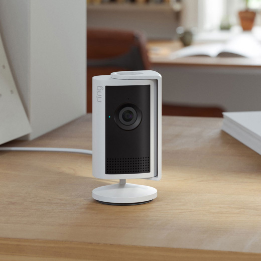 White Ring indoor camera on a wooden tabletop plugged in with a white cord.