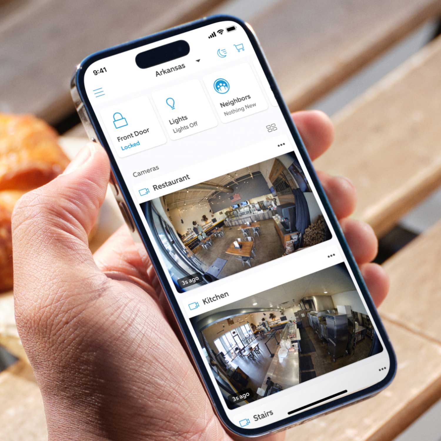 Restaurant Starter Kit (Outdoor Cam Plus Battery + Indoor Cam) - An iPhone in a person's hand with the Ring app open, showing two security camera views of a restaurant.