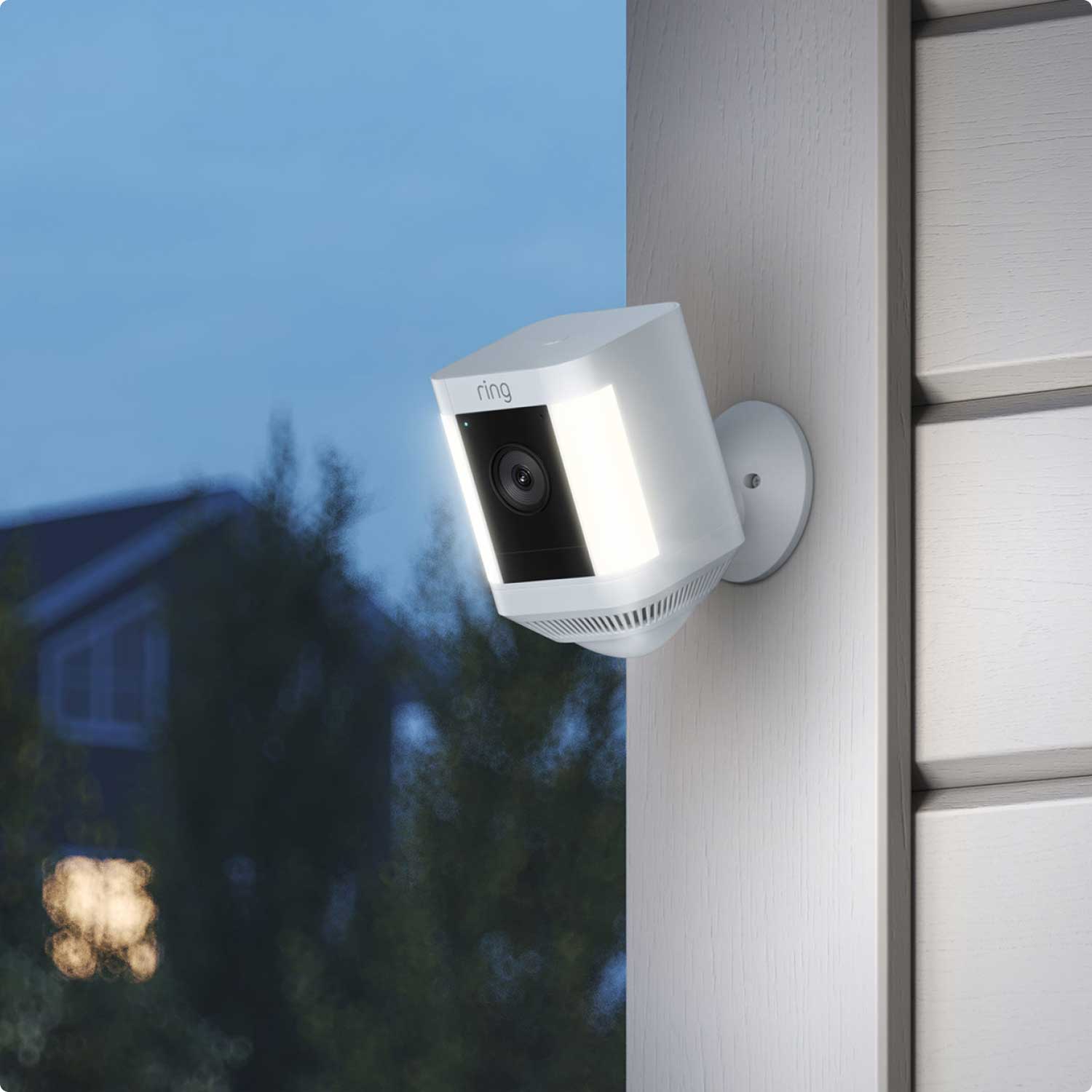 Front and Backyard Basic Kit (Outdoor Cam Plus Battery + Spotlight Cam Plus Battery) - A white Spotlight Cam Plus mounted to corner of house with beige siding, its bright lights turned on in the evening.