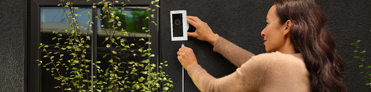 Installing a white Ring security camera on exterior wall near green vines